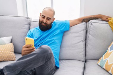 Genç kel adam akıllı telefon kullanıyor. Evdeki koltukta oturuyor.