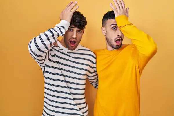 Young hispanic gay couple standing over yellow background surprised with hand on head for mistake, remember error. forgot, bad memory concept.