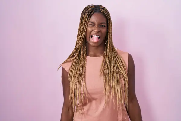 一个有着辫子的非裔美国女人站在粉色的背景上 带着滑稽的表情伸出舌头 情感概念 — 图库照片