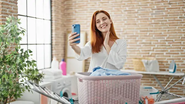 自信を持って笑っている若い赤毛の女性は 洗濯室でスマートフォンでセルフィーを作ります — ストック写真