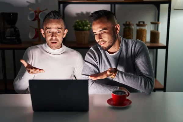 Homosexual couple using computer laptop pointing aside with hands open palms showing copy space, presenting advertisement smiling excited happy