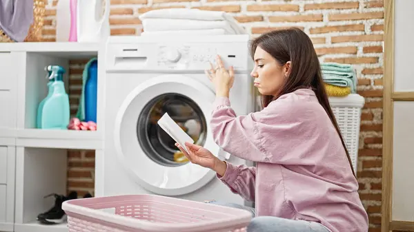 Young beautiful hispanic woman reading instructions to start washing machine at laundry room