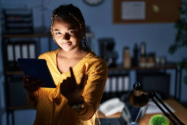 African american woman with braids working at the office at night with tablet beckoning come here gesture with hand inviting welcoming happy and smiling