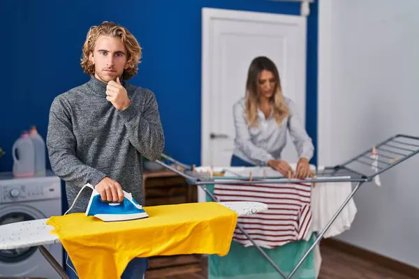 Couple ironing clothes at laundry room serious face thinking about question with hand on chin, thoughtful about confusing idea