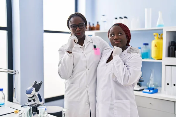 Two african women working at scientist laboratory serious face thinking about question with hand on chin, thoughtful about confusing idea