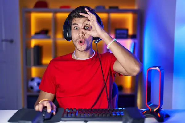 Young hispanic man playing video games doing ok gesture shocked with surprised face, eye looking through fingers. unbelieving expression.