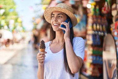 Young hispanic woman tourist talking on the smartphone eating ice cream at street market