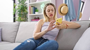 Evdeki kanepede oturan akıllı telefon kullanan genç sarışın kadın.