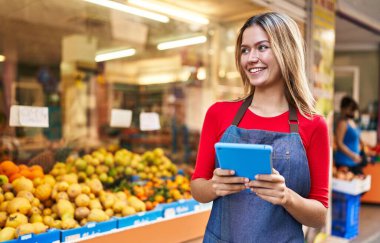 Young hispanic woman shop assistant using touchpad at fruit market