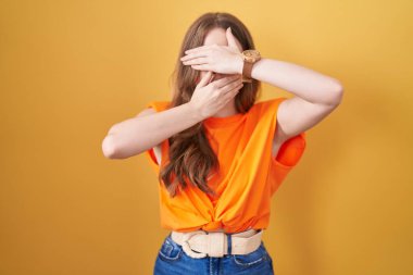 Sarı arka planda dikilen beyaz kadın elleriyle gözlerini ve ağzını kapatıyor, şaşırmış ve şok olmuş. duygu gizleme 