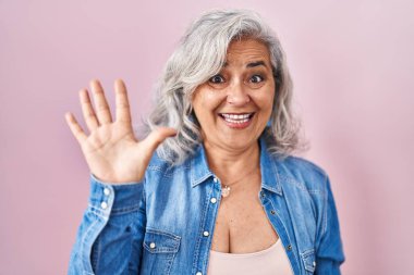 Middle age woman with grey hair standing over pink background showing and pointing up with fingers number five while smiling confident and happy. 