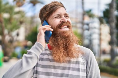 Genç kızıl saçlı, gülümseyen, kendinden emin bir adam caddede akıllı telefondan konuşuyor.