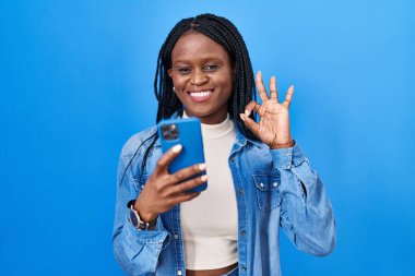 Örgülü Afrikalı kadın akıllı telefon kullanarak mesaj yazıyor parmaklarıyla imza atıyor, gülümseyerek mükemmel bir sembol oluşturuyor. 