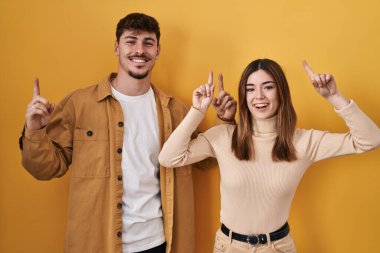 Sarı arka planda dikilen genç İspanyol çift şaşırmış, şaşırmış ve parmaklarıyla işaret ederek kollarını kaldırdı.. 