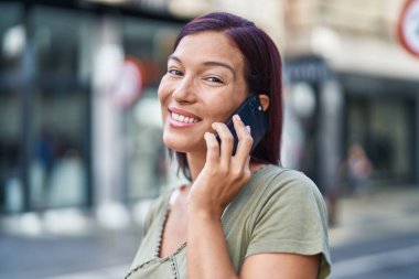 Genç, güzel, İspanyol bir kadın gülümsüyor ve caddede akıllı telefondan konuşuyor.