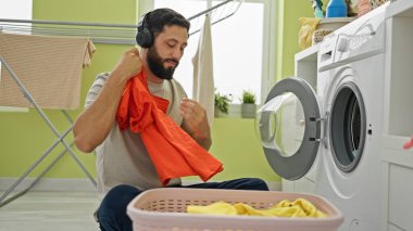 Genç İspanyol adam müzik dinliyor çamaşırhanede giysileri yıkıyor gülümsüyor.