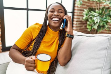 Afro-Amerikalı kadın akıllı telefondan konuşuyor evde kahve içiyor.