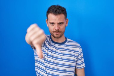 Young hispanic man standing over blue background looking unhappy and angry showing rejection and negative with thumbs down gesture. bad expression. 