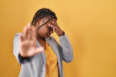 African american woman with braids standing over yellow background covering eyes with hands and doing stop gesture with sad and fear expression. embarrassed and negative concept. 