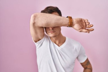 Pembe arka planda duran beyaz bir adam kollarıyla gözlerini kapatıyor, ciddi ve üzgün görünüyor. Görme, saklama ve reddetme konsepti 