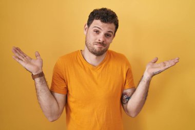 Young hispanic man standing over yellow background clueless and confused expression with arms and hands raised. doubt concept. 