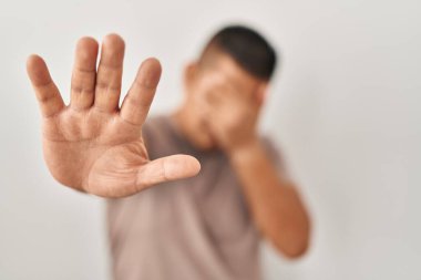 Latin kökenli genç bir adam beyaz arka planda duruyor. Gözleri ellerle kaplıyor ve üzgün ve korku dolu bir ifadeyle jesti durduruyor. utangaç ve olumsuz kavram. 