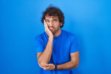 Mavi arka planda duran İspanyol genç adam çapraz kollarla depresyon sorunlarından yorgun ve sıkılmış göründüğünü düşünüyor.. 