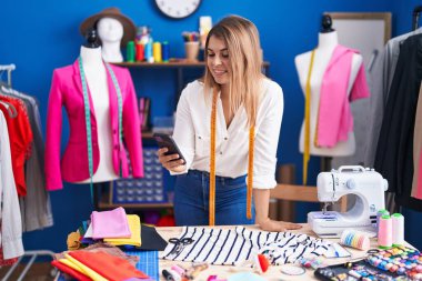 Young woman tailor smiling confident using smartphone at sewing studio