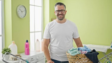 Gri saçlı, gülümseyen bir adam çamaşır odasında çamaşır ipinin yanında duruyor.