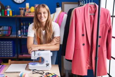 Young blonde girl tailor smiling confident leaning on sewing maching at sewing studio