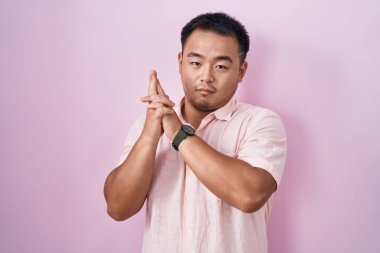 Pembe arka planda duran Çinli genç adam sembolik bir silahla el hareketleriyle cinayet silahlarıyla oynuyor, kızgın surat. 