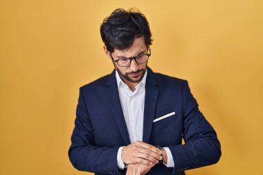 Sarı arka planda duran yakışıklı Latin adam kol saatini kontrol ediyor, rahat ve kendinden emin. 