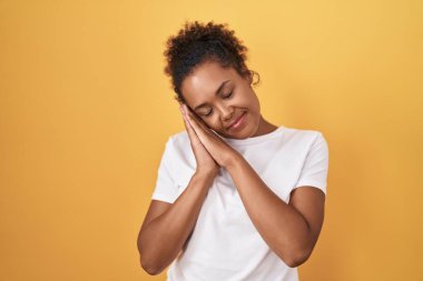 Young hispanic woman with curly hair standing over yellow background sleeping tired dreaming and posing with hands together while smiling with closed eyes. 