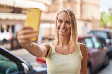 Genç sarışın kadın, kendinden emin gülümsüyor. Sokakta akıllı telefonun yanında özçekim yapıyor.