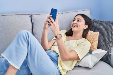 Evdeki kanepede uzanmış akıllı telefon kullanan genç İspanyol kadın.