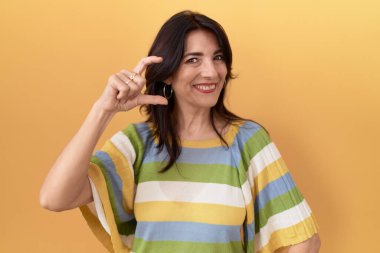 Orta yaşlı İspanyol bir kadın, sarı arka planda durup gülümsüyor ve el hareketleriyle küçük boy işareti yapıyor, parmaklarıyla bakıyor ve kameraya bakıyor. ölçüm kavramı. 