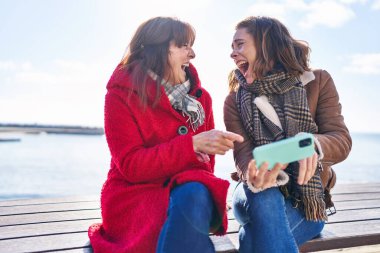 Deniz kenarındaki bankta oturan akıllı telefon kullanan iki kadın anne ve kız.