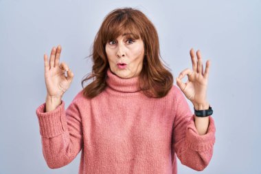 Orta yaşlı İspanyol kadın izole edilmiş arka planda duruyor. Şaşırmış ve şok olmuş görünüyor. Tamam sembolünü parmaklarıyla yapıyor. çılgın ifade 