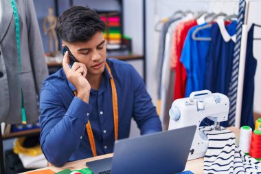 Genç İspanyol terzi elbise fabrikasında dizüstü bilgisayar kullanarak akıllı telefondan konuşuyor.