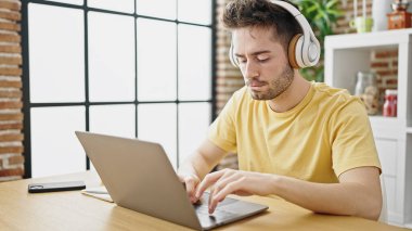 Yemek odasındaki masada dizüstü bilgisayar ve kulaklık kullanan İspanyol genç adam.