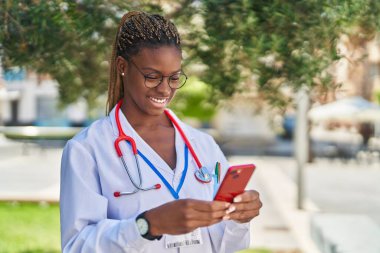 Parkta akıllı telefon kullanarak gülümseyen Afrikalı Amerikalı kadın doktor.