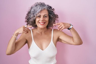 Pembe arka planda duran gri saçlı, neşeli gülümseyen ve parmaklarını ve ağzını gösteren orta yaşlı bir kadın. diş sağlığı kavramı. 