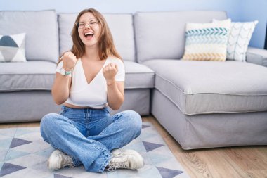 Genç, beyaz bir kadın oturma odasında yerde mutlu ve heyecanlı bir şekilde oturuyor. Kazanan jestler yapıyor. Kolları havada, gülümsüyor ve başarı için haykırıyor. kutlama konsepti. 