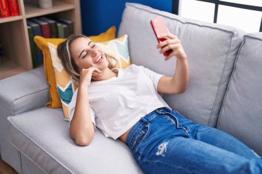 Sarışın genç kadın evdeki kanepede uzanmış video görüşmesi yapıyor.