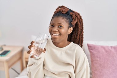 African american woman drinking glass of water sitting on bed at bedroom