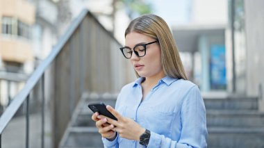 Sarışın genç bir iş kadını sokakta ciddi bir ifadeyle akıllı telefon kullanıyor.