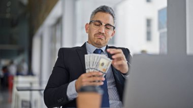 Genç İspanyol iş adamı ofiste dolar sayarak dizüstü bilgisayar kullanıyor.