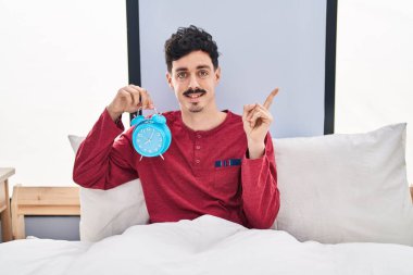 Yatakta alarmlı saat tutan İspanyol adam gülümsüyor. 