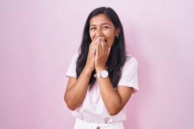 Young hispanic woman standing over pink background laughing and embarrassed giggle covering mouth with hands, gossip and scandal concept 