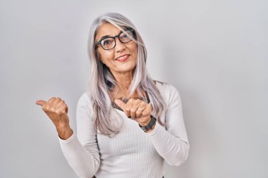 Beyaz arka planda beyaz saçlı, orta yaşlı bir kadın. Arka tarafı işaret ediyor. Baş parmakları havada, kendinden emin gülümsüyor. 